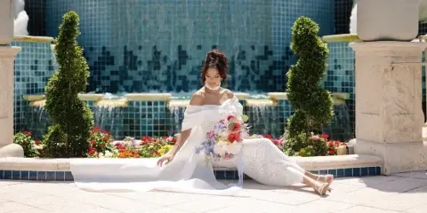 Bride in a white gown seated by a fountain holding a colorful bridal bouquet