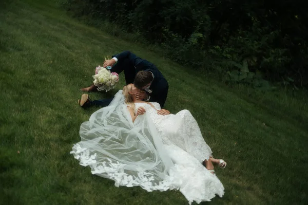 A bride and groom kissing on a grassy lawn, with the bride's white lace dress and veil spread out around them.