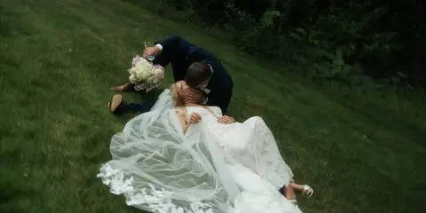 A bride and groom kissing on a grassy lawn, with the bride's white lace dress and veil spread out around them.