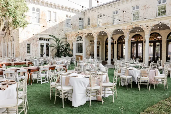 Outdoor wedding reception setup with white tables and chairs on a lawn in front of a historic building with arched windows and string lights.