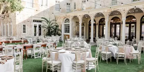 Outdoor wedding reception setup with white tables and chairs on a lawn in front of a historic building with arched windows and string lights.