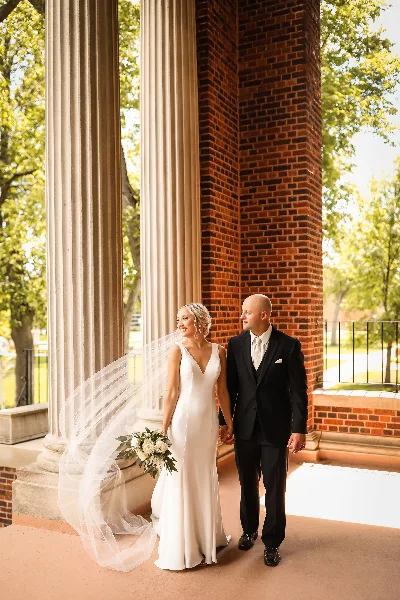 A bride in a white gown and a groom in a black suit walking hand-in-hand under a brick and columned archway.