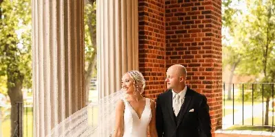 A bride in a white gown and a groom in a black suit walking hand-in-hand under a brick and columned archway.