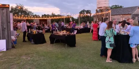 Outdoor evening event at Pioneer Village with string lights, tables, and guests near historic structures.