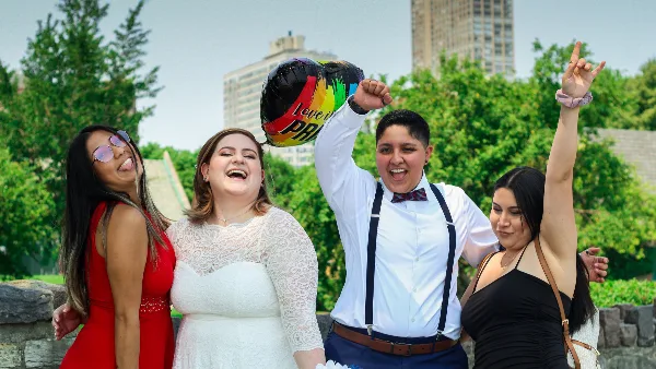 A wedding couple and two guests celebrating outdoors in a park with a rainbow balloon.