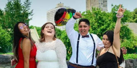 A wedding couple and two guests celebrating outdoors in a park with a rainbow balloon.