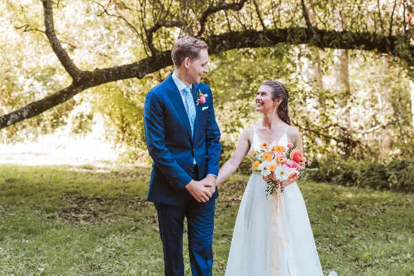 A bride and groom holding hands outdoors, with the bride holding a colorful bouquet of mixed flowers and the groom wearing a matching boutonniere.