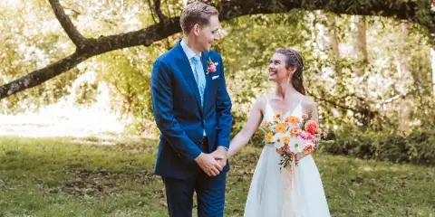A bride and groom holding hands outdoors, with the bride holding a colorful bouquet of mixed flowers and the groom wearing a matching boutonniere.