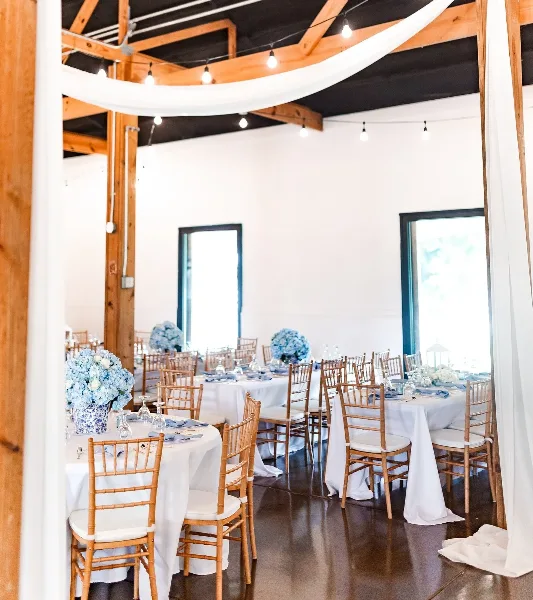 A wedding reception hall with round tables, white linens, blue floral centerpieces, and ceiling drapery.