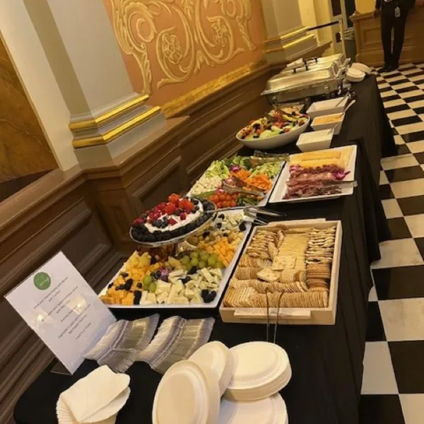 A long buffet table covered with a black tablecloth featuring various food items including a charcuterie board, fruit platters, and chafing dishes.