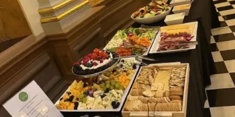 A long buffet table covered with a black tablecloth featuring various food items including a charcuterie board, fruit platters, and chafing dishes.