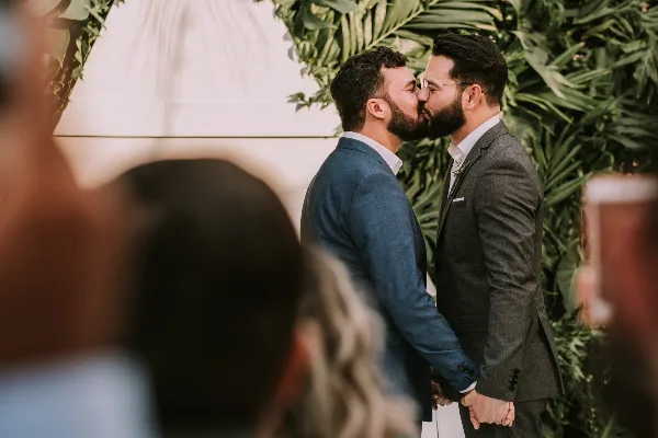 Two men in suits kissing at a wedding altar with a green leafy backdrop and guests watching.