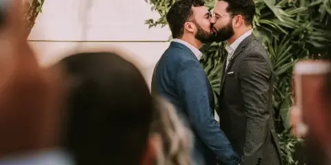 Two men in suits kissing at a wedding altar with a green leafy backdrop and guests watching.