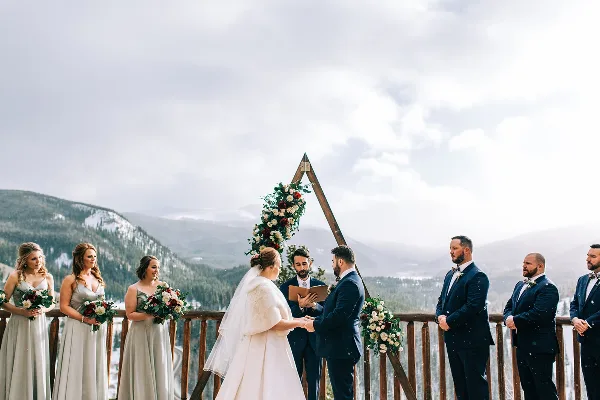 A wedding ceremony taking place on a wooden deck with a mountain backdrop, featuring a bride, groom, officiant, and wedding party.