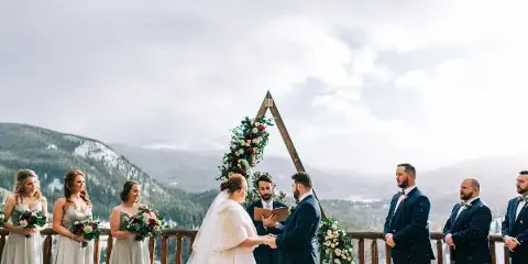 A wedding ceremony taking place on a wooden deck with a mountain backdrop, featuring a bride, groom, officiant, and wedding party.