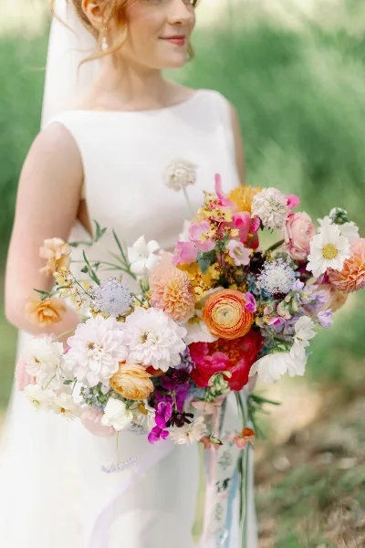 A bride in a white dress holding a vibrant, mixed-color wedding bouquet featuring dahlias, ranunculus, and other seasonal flowers.