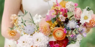 A bride in a white dress holding a vibrant, mixed-color wedding bouquet featuring dahlias, ranunculus, and other seasonal flowers.