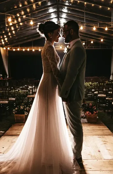 A bride and groom standing face-to-face in a large event tent with string lights and set tables.