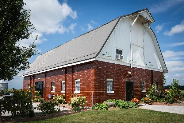 Exterior view of a rustic brick and white barn wedding venue with a metal roof, surrounded by a garden with sunflowers and other plants under a blue sky.