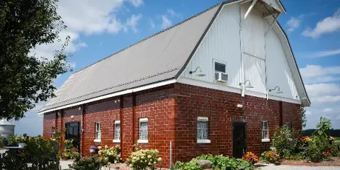 Exterior view of a rustic brick and white barn wedding venue with a metal roof, surrounded by a garden with sunflowers and other plants under a blue sky.