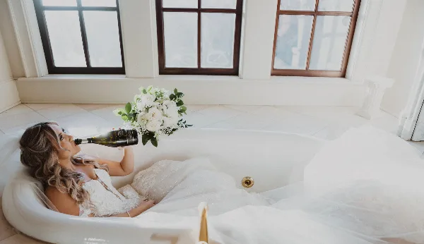 A bride with styled hair and makeup relaxing in a bathtub with a bouquet of white flowers and a bottle of champagne.
