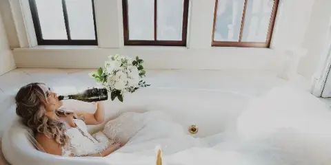 A bride with styled hair and makeup relaxing in a bathtub with a bouquet of white flowers and a bottle of champagne.