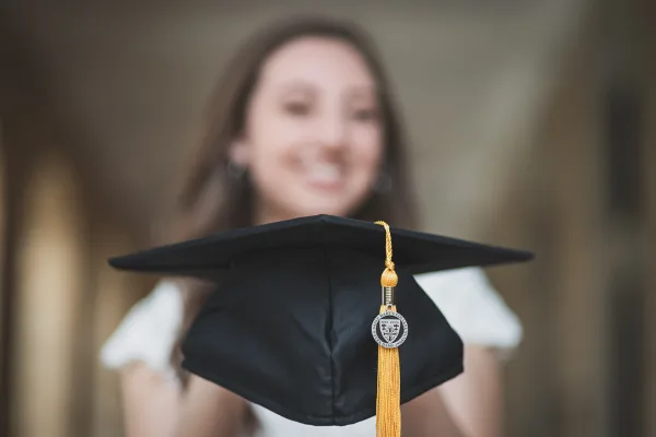 A professional portrait of a smiling young woman holding a black graduation cap with a gold tassel in the foreground.