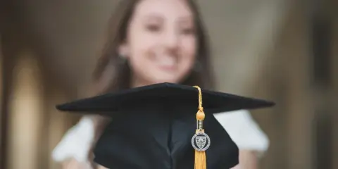 A professional portrait of a smiling young woman holding a black graduation cap with a gold tassel in the foreground.