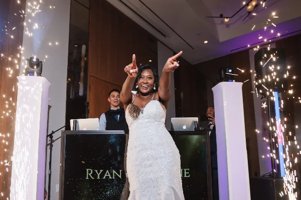 A bride dancing at a wedding reception with a DJ booth and sparkler effects in the background.