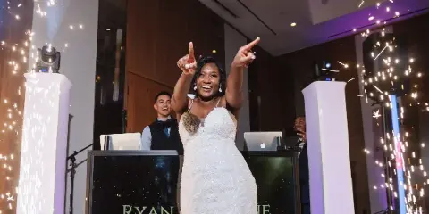 A bride dancing at a wedding reception with a DJ booth and sparkler effects in the background.