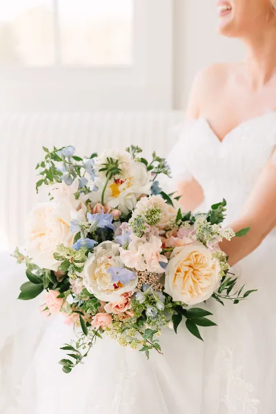 A bride in a white gown holding a large, lush bouquet of white, peach, and blue flowers with greenery.