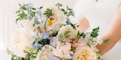 A bride in a white gown holding a large, lush bouquet of white, peach, and blue flowers with greenery.