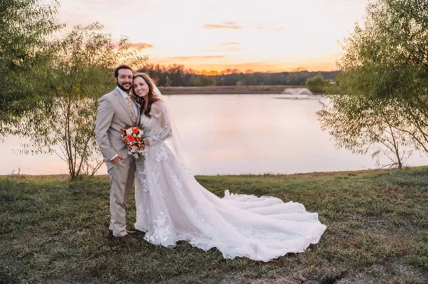 A bride and groom standing on a grassy bank by a lake at sunset.