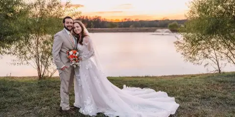 A bride and groom standing on a grassy bank by a lake at sunset.