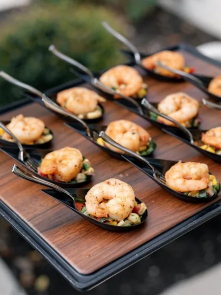 Tray of shrimp appetizers on black serving spoons arranged over a wooden board at an outdoor event.