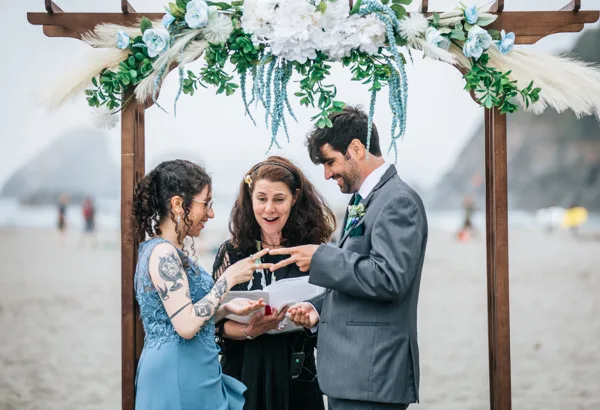 A bride and groom exchanging rings under a floral arch during a beach wedding ceremony.