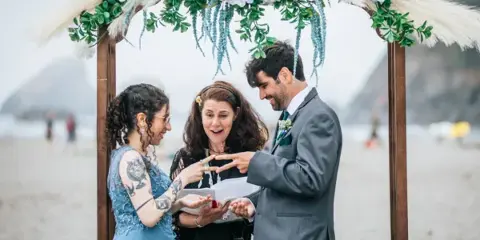 A bride and groom exchanging rings under a floral arch during a beach wedding ceremony.