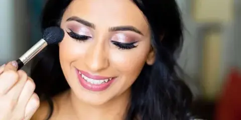 A makeup artist applying eye makeup to a smiling woman with dark hair.