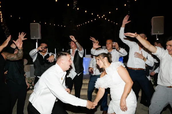 A wedding couple and guests dancing at a reception with a DJ setup in the background.