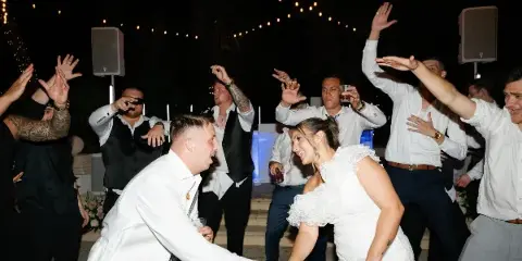 A wedding couple and guests dancing at a reception with a DJ setup in the background.