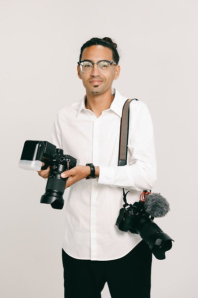 A man in a white shirt holding a professional camera with a flash unit and a second camera with a microphone attached.