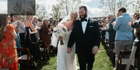 A bride and groom walking down the aisle during an outdoor wedding ceremony with guests seated on either side.