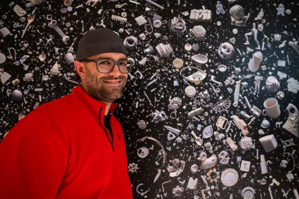 Portrait of Jason Martin, owner of Martin Media Production, wearing a red shirt and cap, standing in front of a wall of 3D printed objects.
