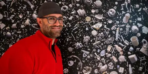 Portrait of Jason Martin, owner of Martin Media Production, wearing a red shirt and cap, standing in front of a wall of 3D printed objects.