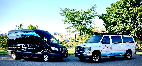 Two branded passenger vans, one black and one white, parked outdoors with The Local Transport logos and contact information on the sides.