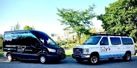 Two branded passenger vans, one black and one white, parked outdoors with The Local Transport logos and contact information on the sides.