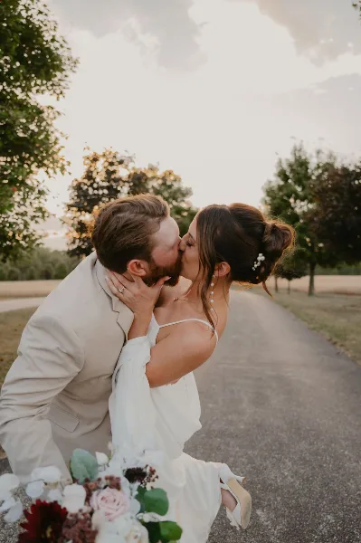 A bride and groom sharing a kiss outdoors on a paved path with trees in the background.