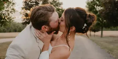 A bride and groom sharing a kiss outdoors on a paved path with trees in the background.
