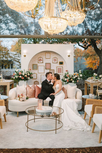 A professionally designed wedding reception area featuring a couple on a white sofa, surrounded by floral arrangements, a decorative arched backdrop, and crystal chandeliers.