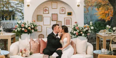 A professionally designed wedding reception area featuring a couple on a white sofa, surrounded by floral arrangements, a decorative arched backdrop, and crystal chandeliers.